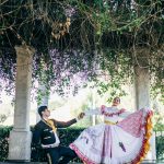 A couple performs a traditional Mexican dance in colorful outfits under a lush pergola.