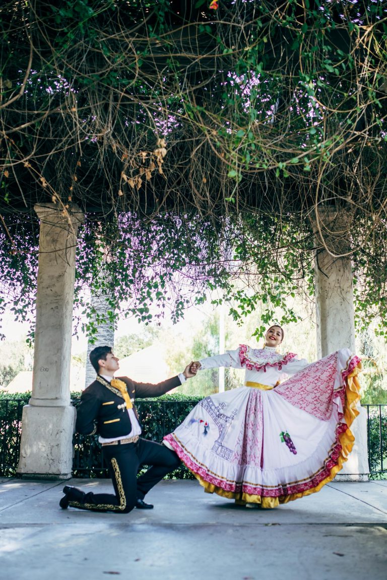 A couple performs a traditional Mexican dance in colorful outfits under a lush pergola.