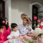 A joyful family gathering featuring a man in a sombrero, surrounded by flowers and food at a festive table.