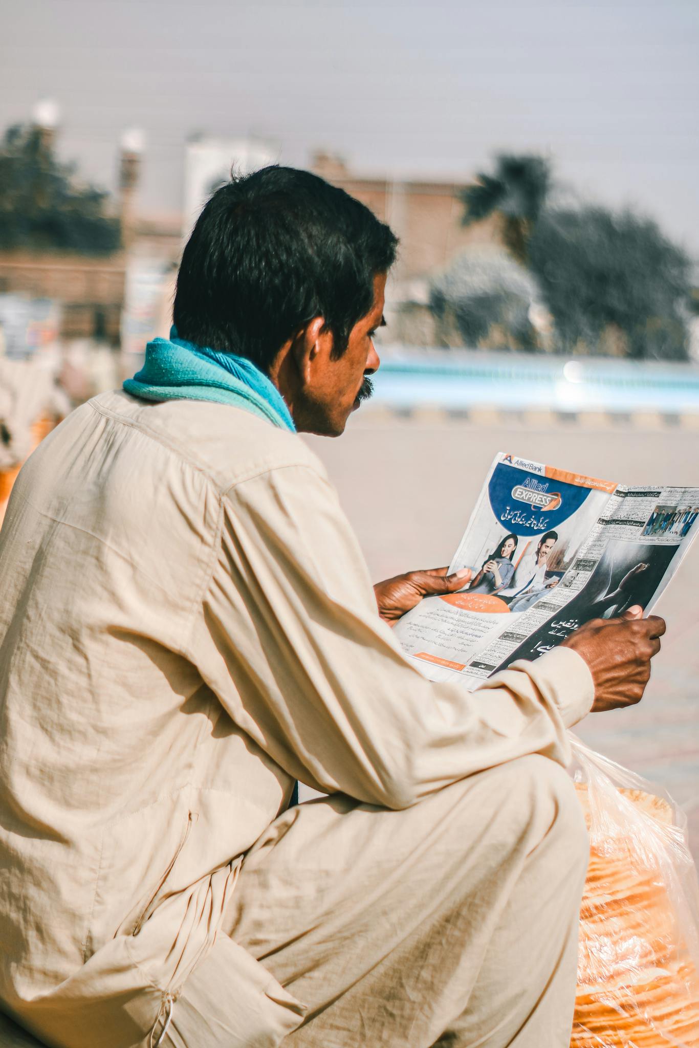 A man in traditional attire reads a newspaper in an outdoor setting in Multan, Pakistan.