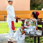 Man and woman in traditional Yucatecan clothing interacting in a vibrant park setting.