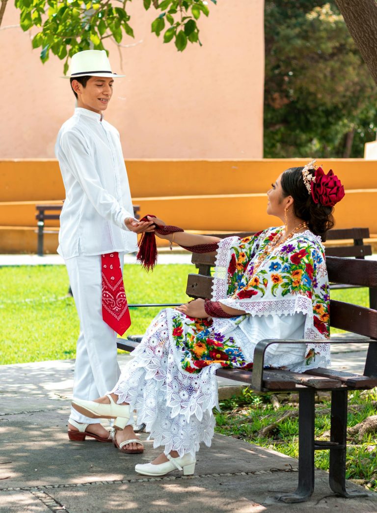 Man and woman in traditional Yucatecan clothing interacting in a vibrant park setting.